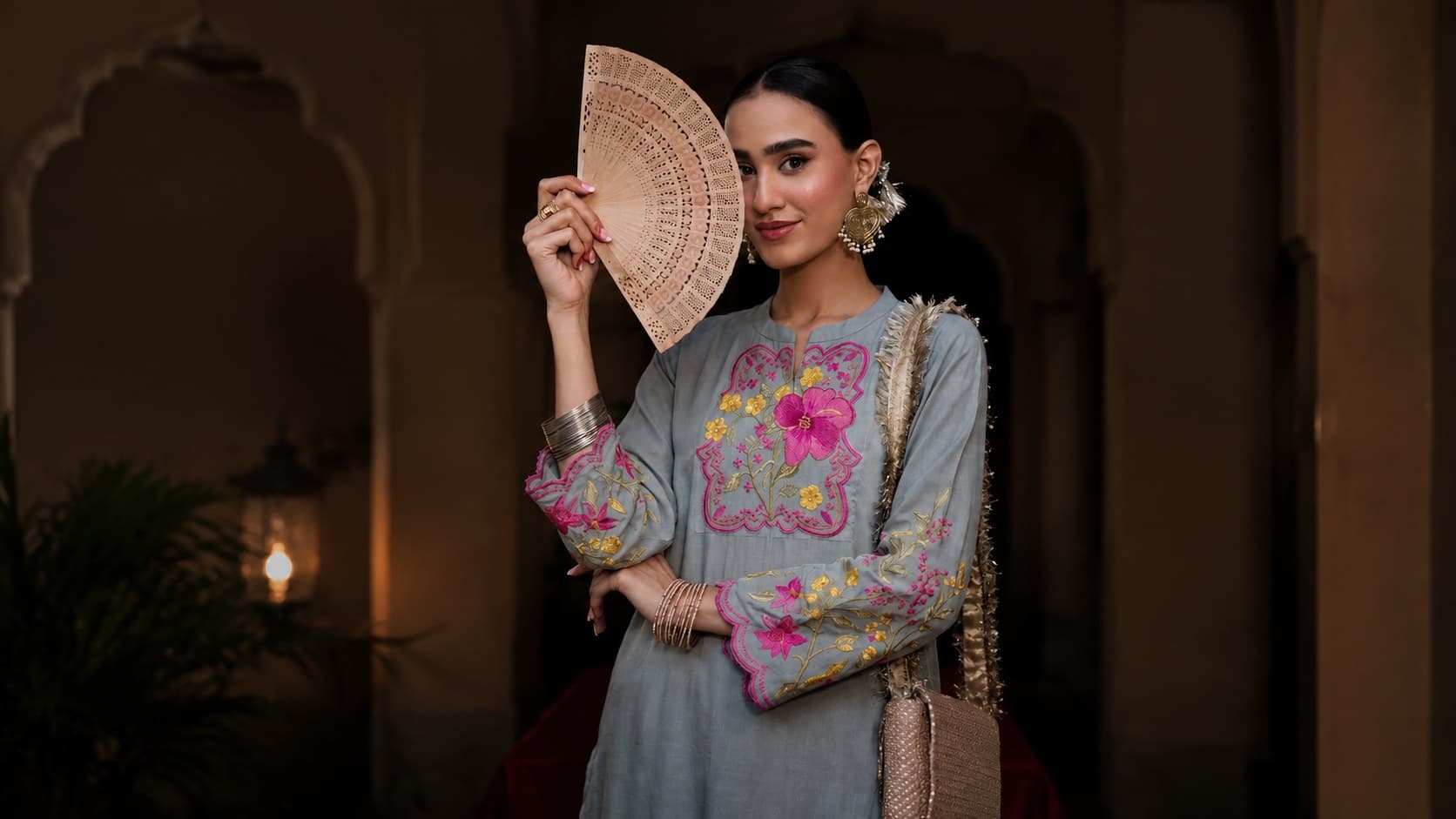 Grey-blue kurta with pink and gold floral embroidery, styled with a carved wooden fan in a lamplit haveli archway